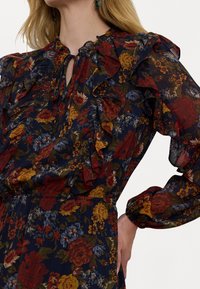 Ziedu blūze tumši zilā krāsā, ar frillēm, garām piedurknēm un atslēgvārsta izgriezumu. Rakstā ietilpst sarkani, dzelteni un zili ziedi.