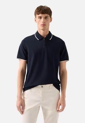 Jonge man draagt een marineblauw poloshirt met korte mouwen en witte bies en beige broek, staand tegen een effen lichte achtergrond.