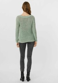 Maglione lavorato a maglia verde chiaro con una vestibilità ampia e maniche lunghe, abbinato a pantaloni slim-fit grigio scuro e stivali neri alla caviglia.