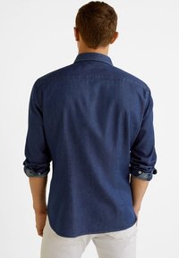 Ciemna koszula z długim rękawem z denimu, z podwiniętymi mankietami i kołnierzykiem na guziki. Charakteryzuje się gładką teksturą i dopasowanym krojem, widziana od tyłu.