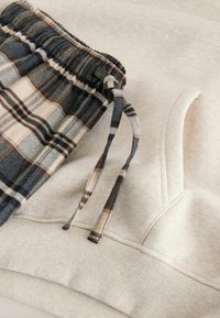 Hanorac bej cu buzunar frontal și tiv cu nervuri, asociat cu pantaloni gri cu model carouri, dotati cu talie elastică.