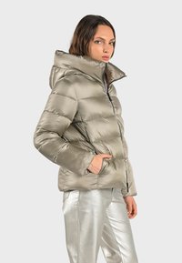 Polstret puffer-jakke i metallisk olivengrøn med hætte, med zip-lukning og sidelommer. Bæres med sølvteksturerede bukser.