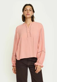Rosa Bluse mit rundem Halsausschnitt, Henley-Knopfleiste und langen Ärmeln. Sie hat gerüschte Bündchen und subtile Nähte entlang der Nähte.
