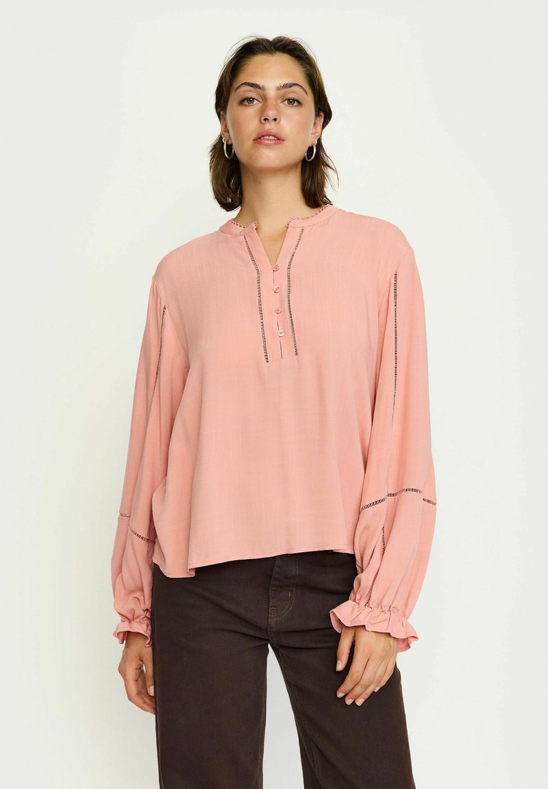Rosa Bluse mit rundem Halsausschnitt, Henley-Knopfleiste und langen Ärmeln. Sie hat gerüschte Bündchen und subtile Nähte entlang der Nähte.