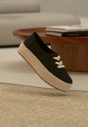 Czarny trampki wsuwany z grubą, plecioną beżową podeszwą na platformie i białymi sznurowadłami, unoszący się nad teksturowanym beżowym dywanem obok drewnianego stołu z książkami.