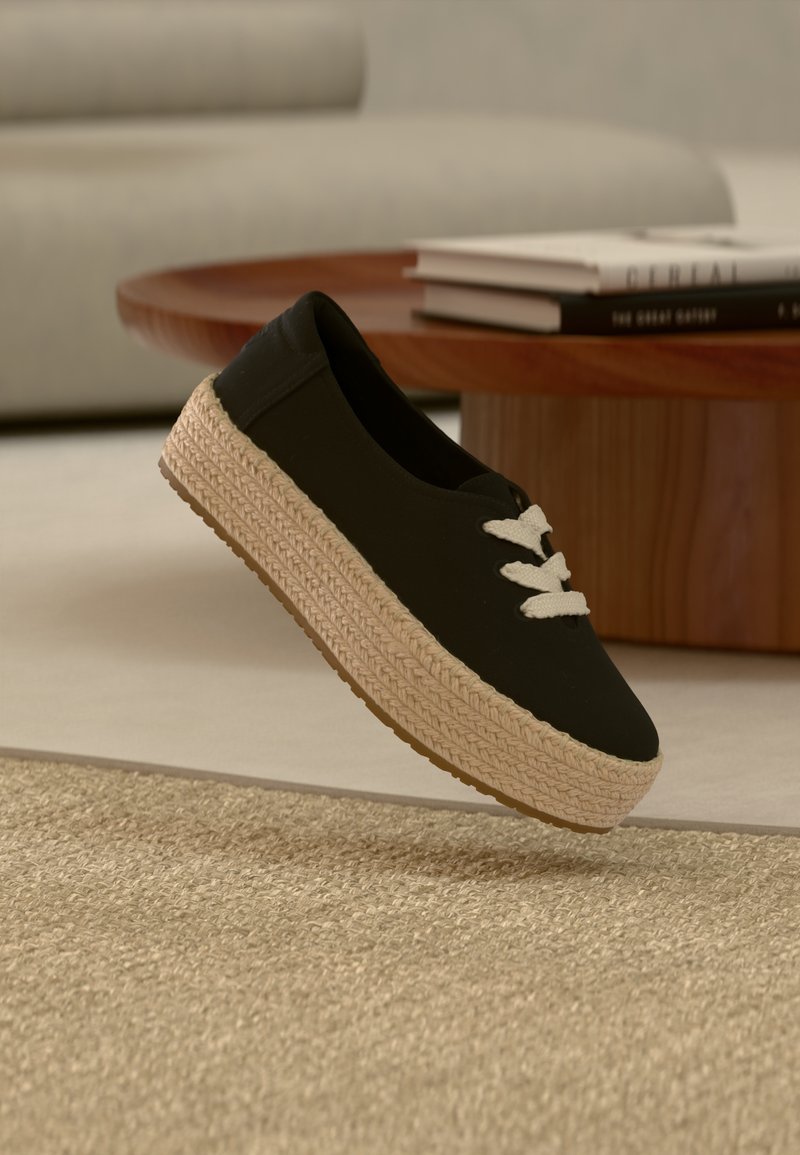 Μαύρο πάνινο παπούτσι χωρίς κορδόνια με χοντρή πλεκτή μπεζ σόλα πλατφόρμας και λευκά κορδόνια, να αιωρείται πάνω από υφασμένο μπεζ χαλί κοντά σε ξύλινο τραπέζι με βιβλία.