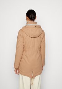 Béžová zateplená parka s kapucí, stahováním v pase a límcem s kožešinou. Má rozdělený zadní díl a dlouhé rukávy s manžetami.