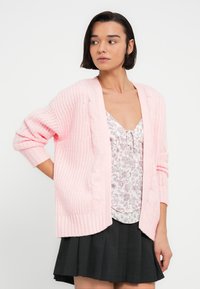 Ανοιχτό ροζ πλεκτή ζακέτα με υφή, που διαθέτει χοντρό πλέξιμο σε σχέδιο καλωδίου κατά μήκος των άκρων και ριμπ μανσέτες. Φορεμένη πάνω από ένα τοπ με λουλούδια.