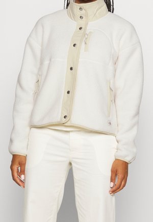 Mujer con chaqueta de forro polar color crema con botones a presión, bolsillos en el pecho y laterales con cremallera, combinada con pantalones de color claro.