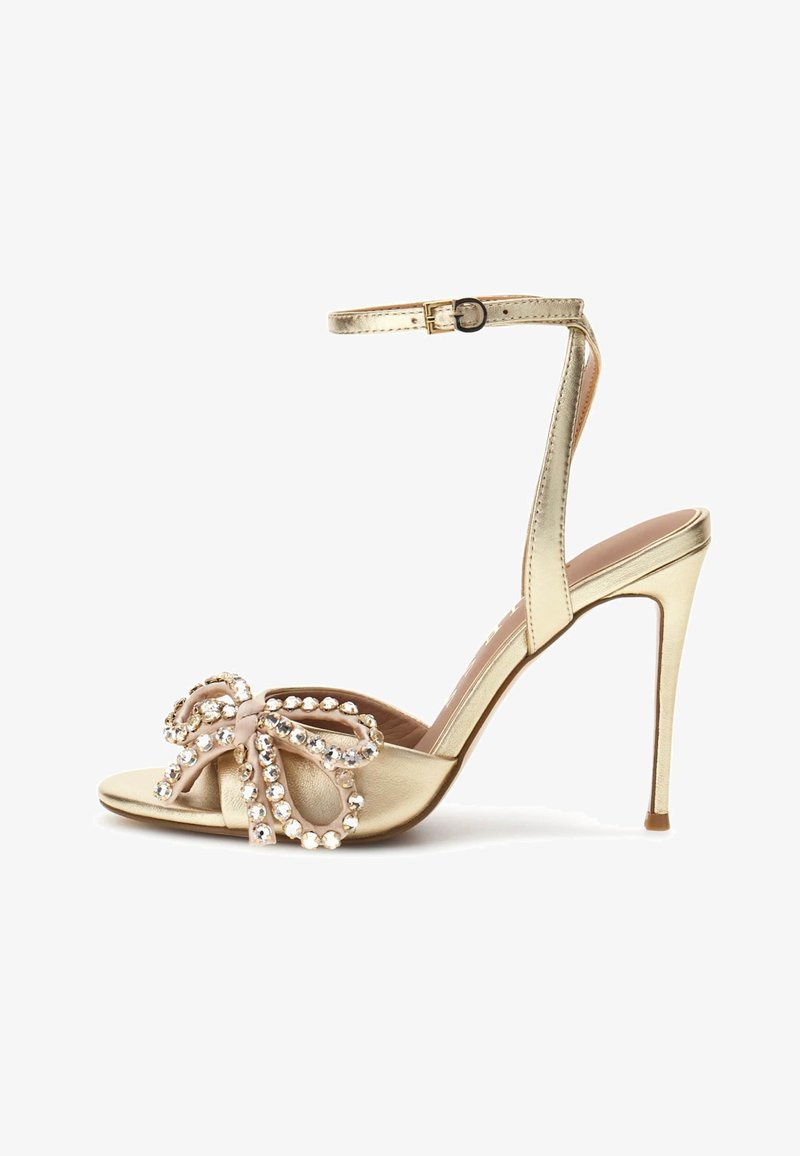 Goldfarbene High-Heel-Sandalen mit Knöchelriemen und offenem Zehenbereich, verziert mit einer dekorativen Schleife, die mit funkelnden Kristallen geschmückt ist.
