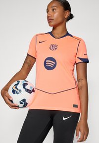 Nike Barcelona-shirt in felrood met marineblauwe accenten en logo. Glad weefsel, korte mouwen, vergezeld van een witte en blauwe voetbal.