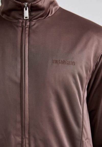 Brun zip-up jakke laget av glatt stoff, med høy krage, subtil tekstur og brodert logo på brystet.