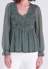 Blouse à motifs verts et crème avec manches longues et transparentes, taille froncée smockée et encolure en V. Tissu texturé avec une coupe fluide.