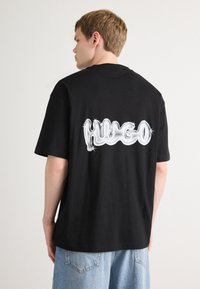 Černé bavlněné tričko s velkým, lesklým bílým logem "Hugo" tištěným na zadní straně. Volný střih, krátké rukávy a kulatý výstřih.
