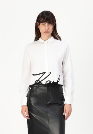 Camicia bianca button-up con colletto classico, abbinata a una gonna a matita nera in pelle con tasche e una texture liscia.