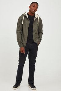 Olivengrøn hættetrøje med lynlås og lysegrå foring, med snoretræk og to forlommer, kombineret med sorte jeans og sorte sneakers.