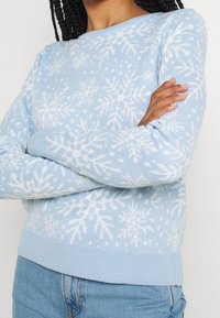 Hellblauer Pullover mit weißen Schneeflockenmuster; verfügt über einen runden Ausschnitt und einen gerippten Saum; weiches Strickmaterial mit langen Ärmeln.