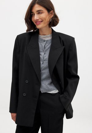 Zwarte dubbelzijdige blazer met brede revers, gestructureerde pasvorm, textuurstof en twee voorsluitingen, gelaagd over een grijze cardigan.