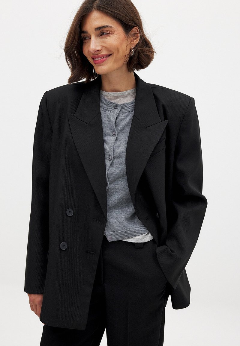 Zwarte dubbelzijdige blazer met brede revers, gestructureerde pasvorm, textuurstof en twee voorsluitingen, gelaagd over een grijze cardigan.