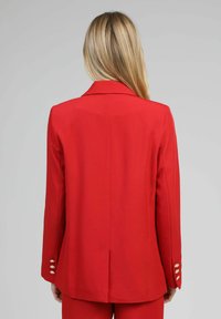 Personne blonde portant un blazer rouge sur mesure et un pantalon rouge assorti, vue de dos sur un fond gris uni.
