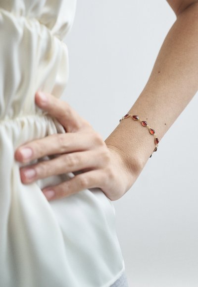 Mâna unei femei odihnindu-se pe talie, purtând o brățară de aur cu pietre în formă de lacrimă roșie și o bluză crem, plisată.