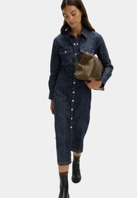 Denimshirtklänning i mörkblått, med knapplås fram, bröstfickor, långa ärmar och midi-längd. Matchad med en brun läderväska.