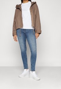 Persona con chaqueta con capucha marrón, camiseta blanca, jeans ajustados azules y zapatillas blancas, de pie contra un fondo liso y claro.