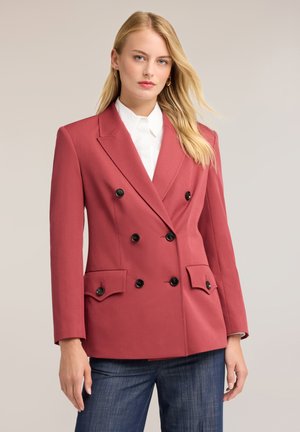 Femme aux longs cheveux blonds portant un blazer rouge à double boutonnage, une chemise blanche et un jean bleu foncé, debout devant un fond clair uni.