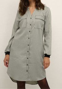 Abito a camicia in fantasia a scacchi bianco e nero, con tasche sul petto, maniche lunghe con polsini neri e orlo leggermente curvato.