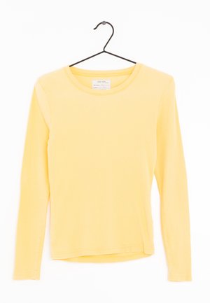 Chemise jaune à manches longues côtelée avec encolure ronde, suspendue à un cintre noir sur fond blanc.