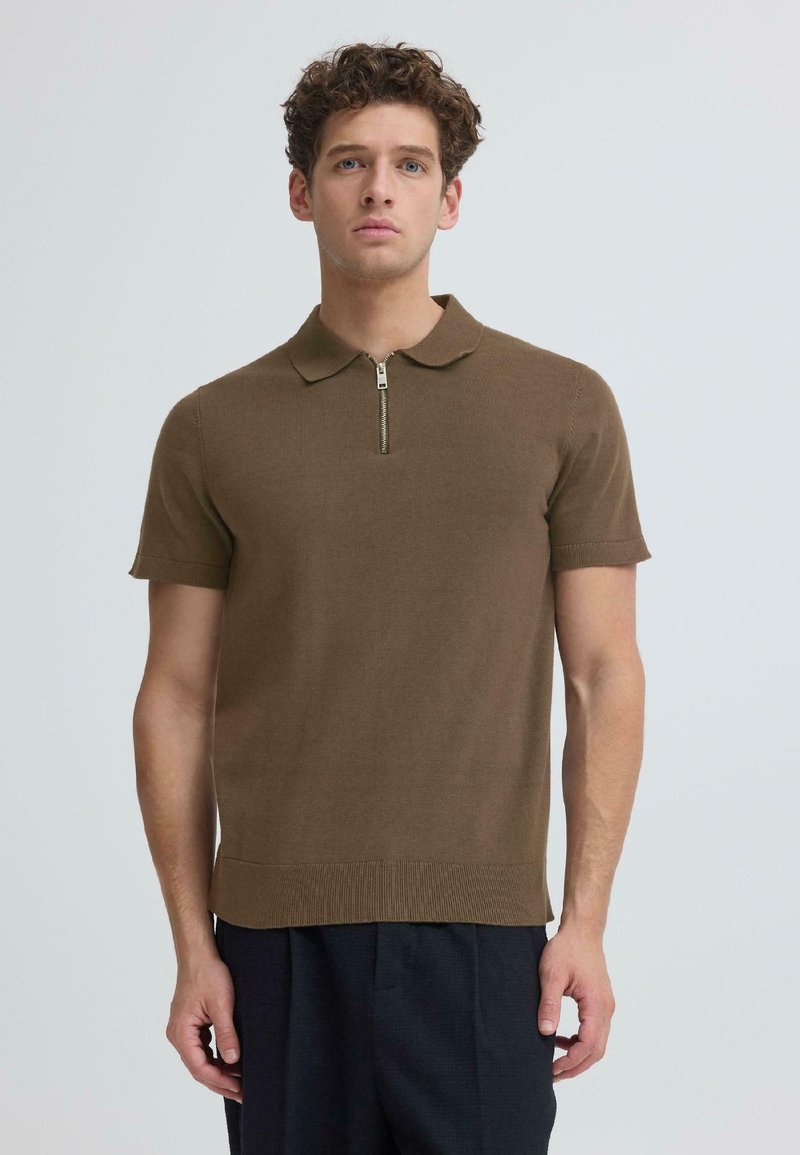 Jeune homme aux cheveux bouclés portant un polo marron à manches courtes avec une fermeture éclair à l'avant et un pantalon sombre, se tenant devant un fond uni.