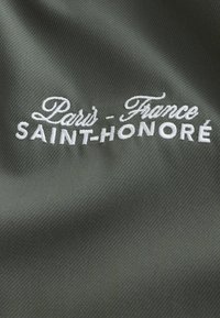 Texte brodé blanc « Paris - France SAINT-HONORÉ » sur tissu vert foncé avec une subtile texture diagonale tissée.