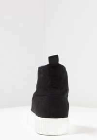 Sort ankelstøvle i lærred med en hvid gummisål, der har en løkke bagpå og syningsdetaljer langs sømmene.