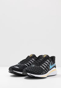 Scarpe da ginnastica nere con suola bianca e gialla, caratterizzate da un logo blu e un tessuto a motivo, esposte su uno sfondo bianco semplice.
