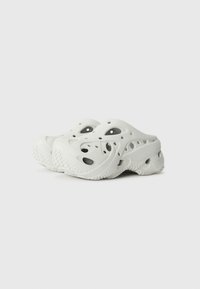 White molded clogs with multiple ventilation holes, thick soles, and a wavy texture pattern on the sides and bottom for grip.