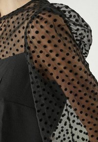 Haut noir avec manches en organza à pois transparents. Les manches sont texturées avec des motifs de points noirs, créant un effet de superposition.