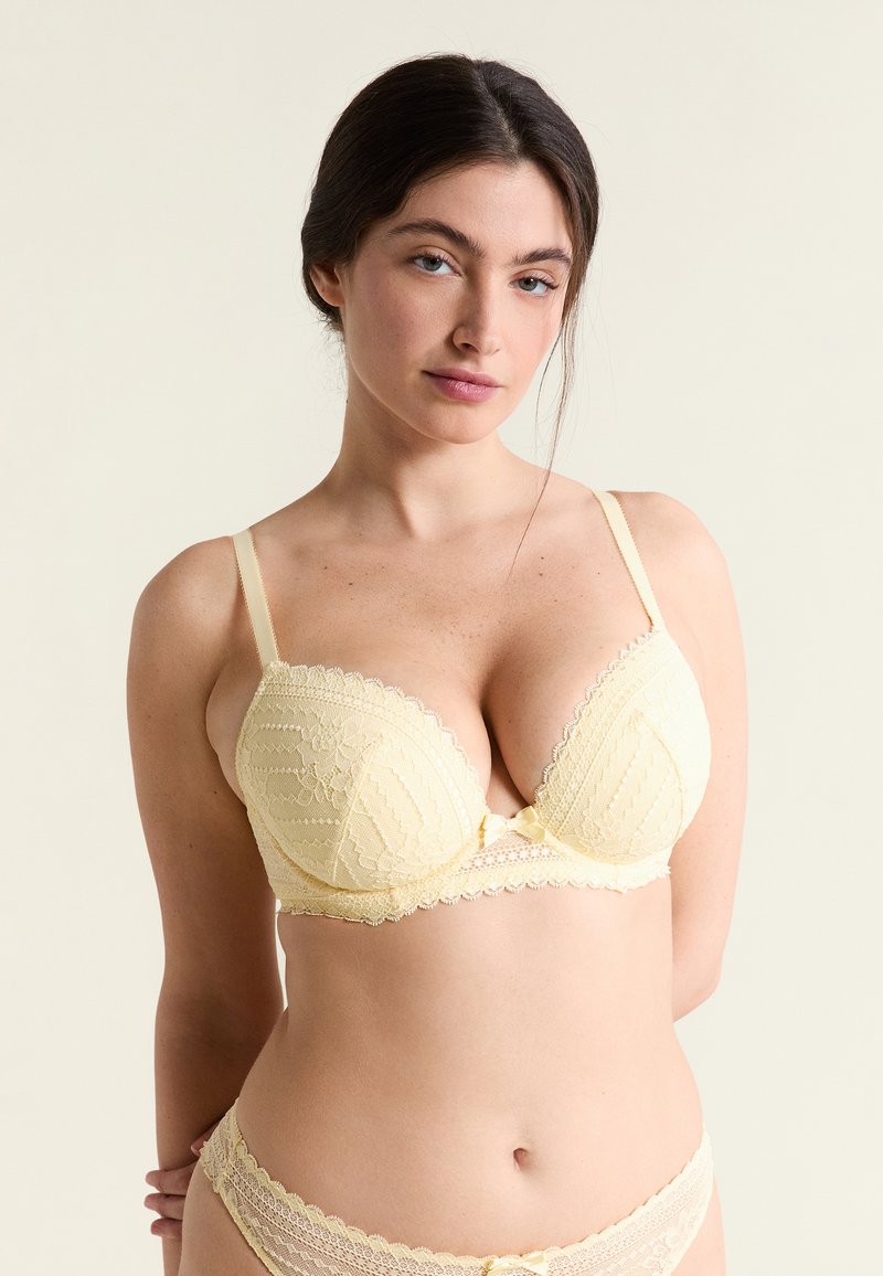 Mujer de cabello oscuro vistiendo un sujetador de encaje amarillo claro y bragas a juego, de pie contra un fondo claro y liso, con las manos detrás de la espalda.