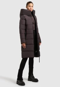 Lange donkerbruine pufferjas met een hoge kraag, voorzien van gestikte delen en zijritsen, gecombineerd met zwarte slim fit broeken en stevige zwarte laarzen.