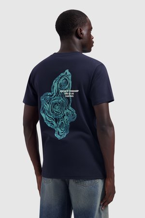Man wearing a navy blue t-shirt with a turquoise tree ring design and white text reading "Rooted by Friendship Tree of Life Pure Path" on the back.