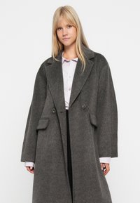 Grijze wollen trenchcoat met een gesneden revers, dubbele knoopsluiting en twee voorkruiszakken. De stof heeft een zachte textuur.