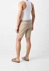 Pantaloni beige con orlo arrotolato, realizzati in tessuto leggero. Cintura intrecciata e due tasche sul retro. Modello che indossa una canotta bianca e sandali.