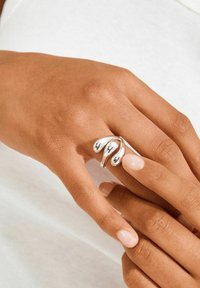 Hõberõngas keeruka disainiga, millel on kaks tilgakujulist aktsenti, kantud käel loodusliku küünelakiga. Tekstuuriga nahataust.