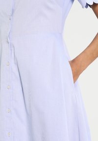 Ljusblå skjortklänning i strukturvävt tyg, med korta ärmar och sidofickor. Har en figurnära överdel och en utsvängd kjoldesign.