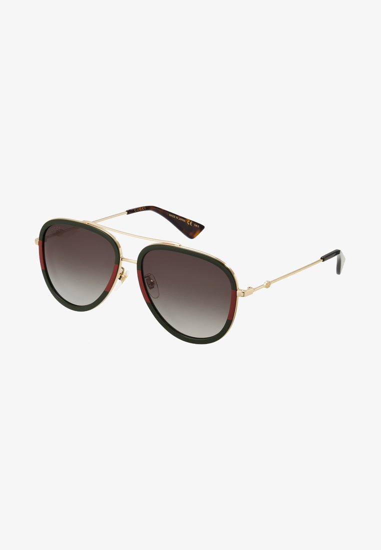 Des lunettes de soleil de style aviateur avec une monture en métal doré, des accents verts et rouges, des verres teintés dégradés foncés et une forme géométrique définie.