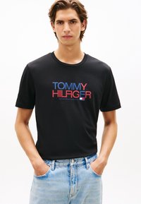 Μαύρο βαμβακερό T-shirt με το λογότυπο "TOMMY HILFIGER" σε κόκκινο και μπλε. Κοντά μανίκια, στρογγυλή λαιμόκοψη και κανονική εφαρμογή. Φοριέται με ανοιχτόχρωμο μπλε τζιν.