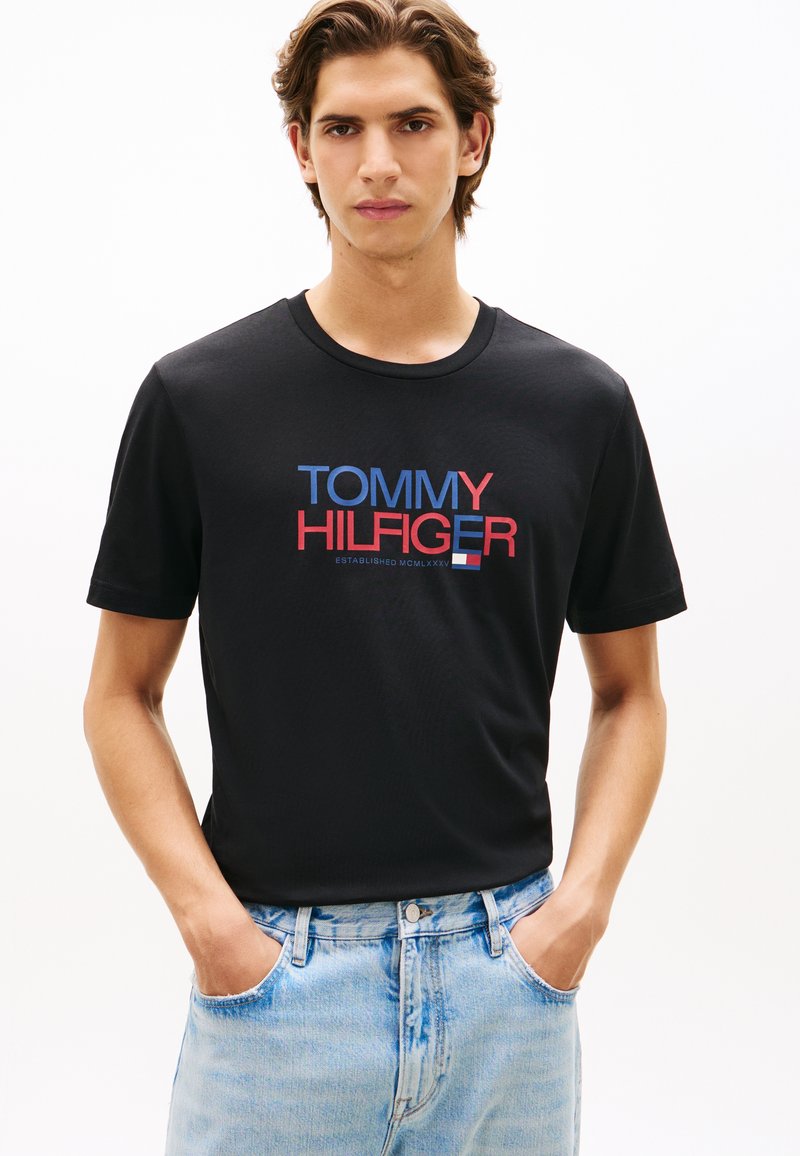 Μαύρο βαμβακερό T-shirt με το λογότυπο "TOMMY HILFIGER" σε κόκκινο και μπλε. Κοντά μανίκια, στρογγυλή λαιμόκοψη και κανονική εφαρμογή. Φοριέται με ανοιχτόχρωμο μπλε τζιν.