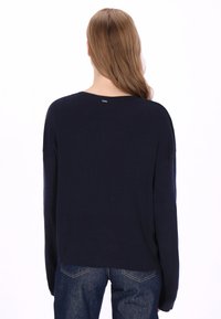 Marineblauwe geribde trui met een ronde hals en een losse pasvorm. Heeft een klein metalen label aan de achterkant. Draagt blauw spijkerbroek.