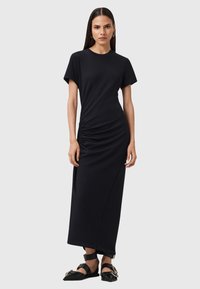 Maxi dress nera a maniche corte, con dettaglio arricciato sul lato, texture liscia e silhouette dritta. Accentuata da sandali.
