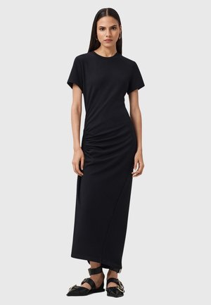 Robe maxi noire en jersey à manches courtes, présentant un détail froncé sur le côté, une texture lisse et une silhouette droite. Accentuée par des sandales.