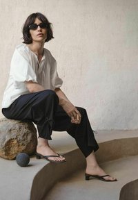 Vrouw met zwarte zonnebril, witte blouse en zwarte broek, zittend op een rots naast een rond zwart object op gebogen betonnen traptreden.